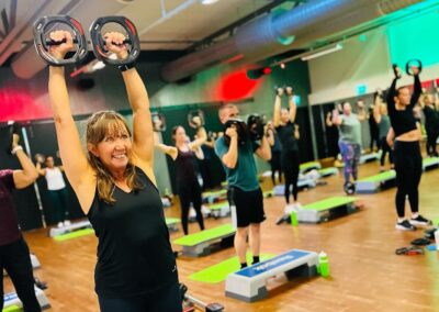 Bilden visar en livlig gruppträningsklass där en kvinna i förgrunden ler och lyfter två kettlebells över huvudet. I bakgrunden syns andra deltagare som också utför övningen, alla fokuserade på sin träning. Rummet är upplyst med färgglada ljus som ger en energisk atmosfär. Träningen verkar vara intensiv men samtidigt njutbar, vilket återspeglas i deltagarnas positiva energi.